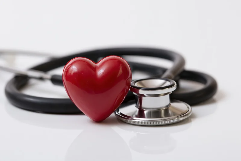 Black stethoscope coiled around small red heart figurine on white reflective surface