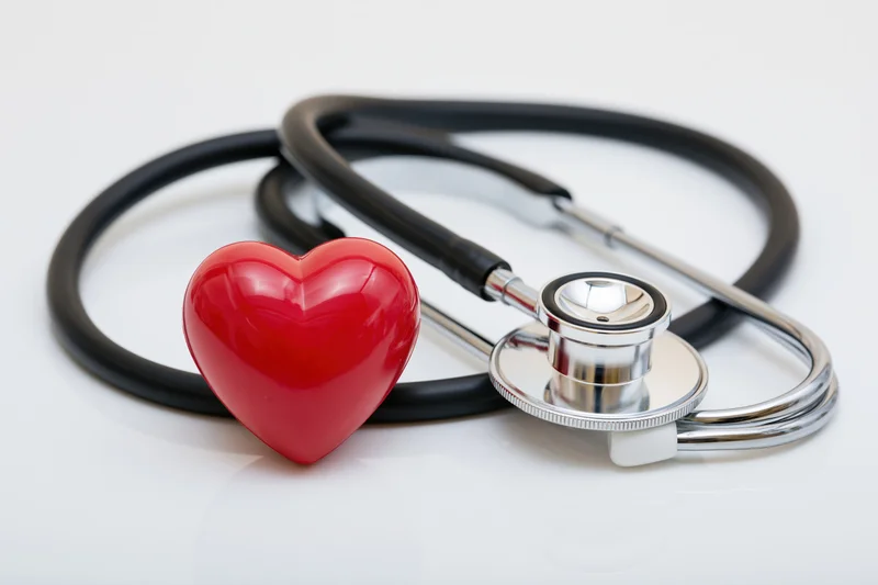 Black stethoscope coiled around a small red heart figurine on white surface