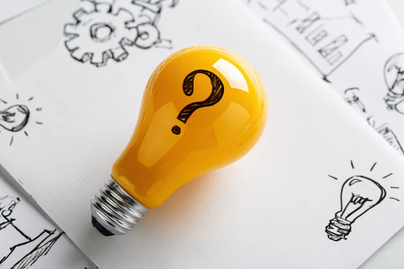 Yellow lightbulb with question mark resting on paper with hand-drawn sketches of ideas and diagrams