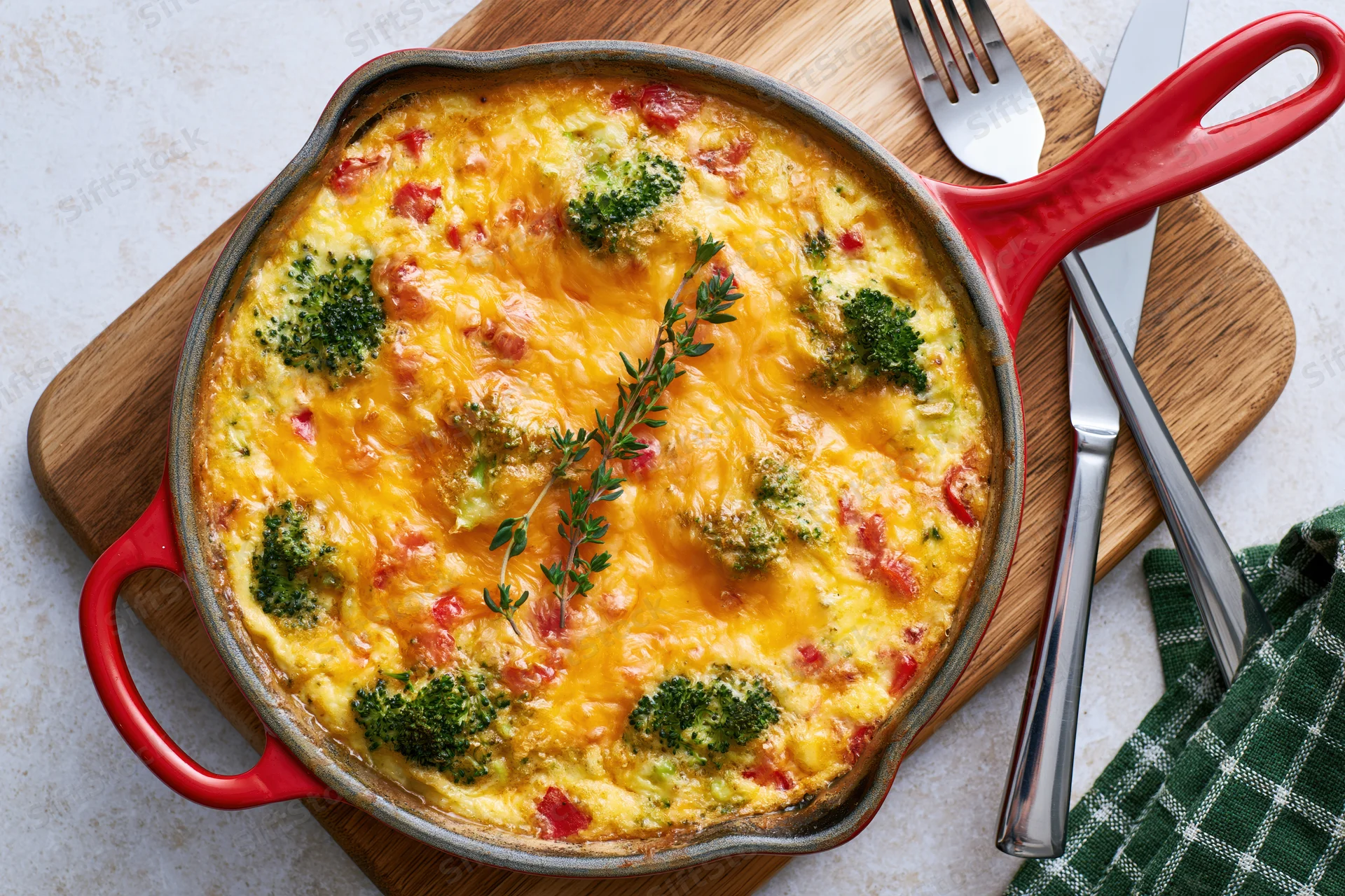 Baked broccoli and red pepper frittata in a red-handled skillet on a wooden board with cutlery and a checkered napkin