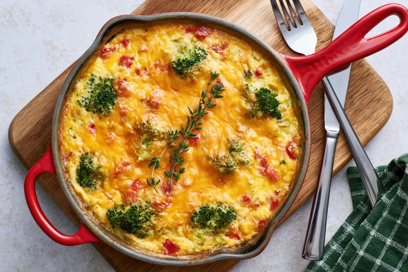 Baked broccoli and red pepper frittata in a red-handled skillet on a wooden board with cutlery and a checkered napkin
