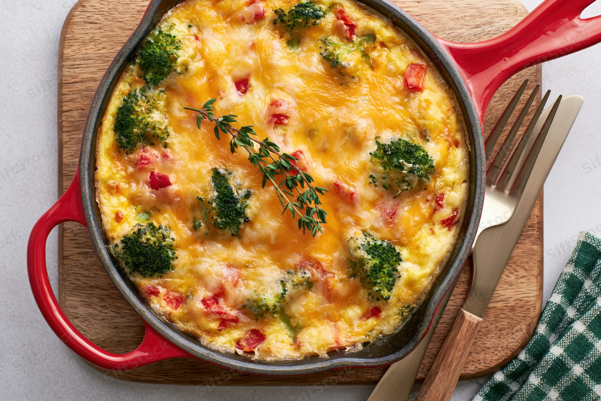 Broccoli and red pepper frittata baked in a red-handled skillet on a wooden cutting board with fork and knife beside it