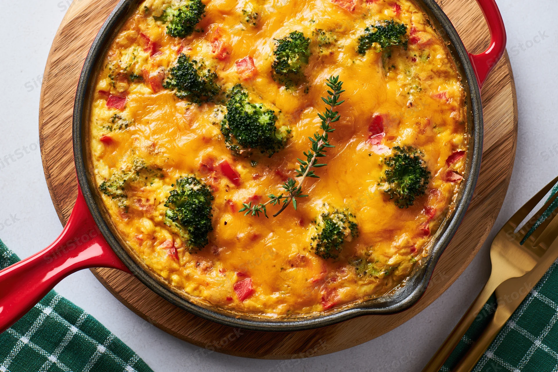Broccoli and red pepper frittata baked in a red-handled skillet on a wooden tray with a fork and green checkered napkin