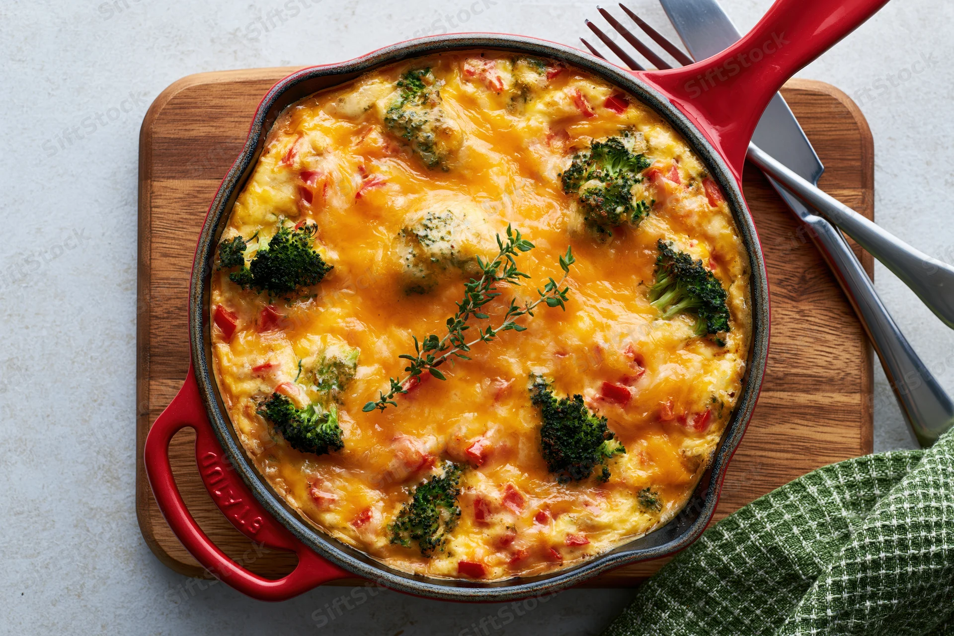 Baked broccoli and red pepper frittata in red cast iron skillet on wooden cutting board with fork and knife nearby