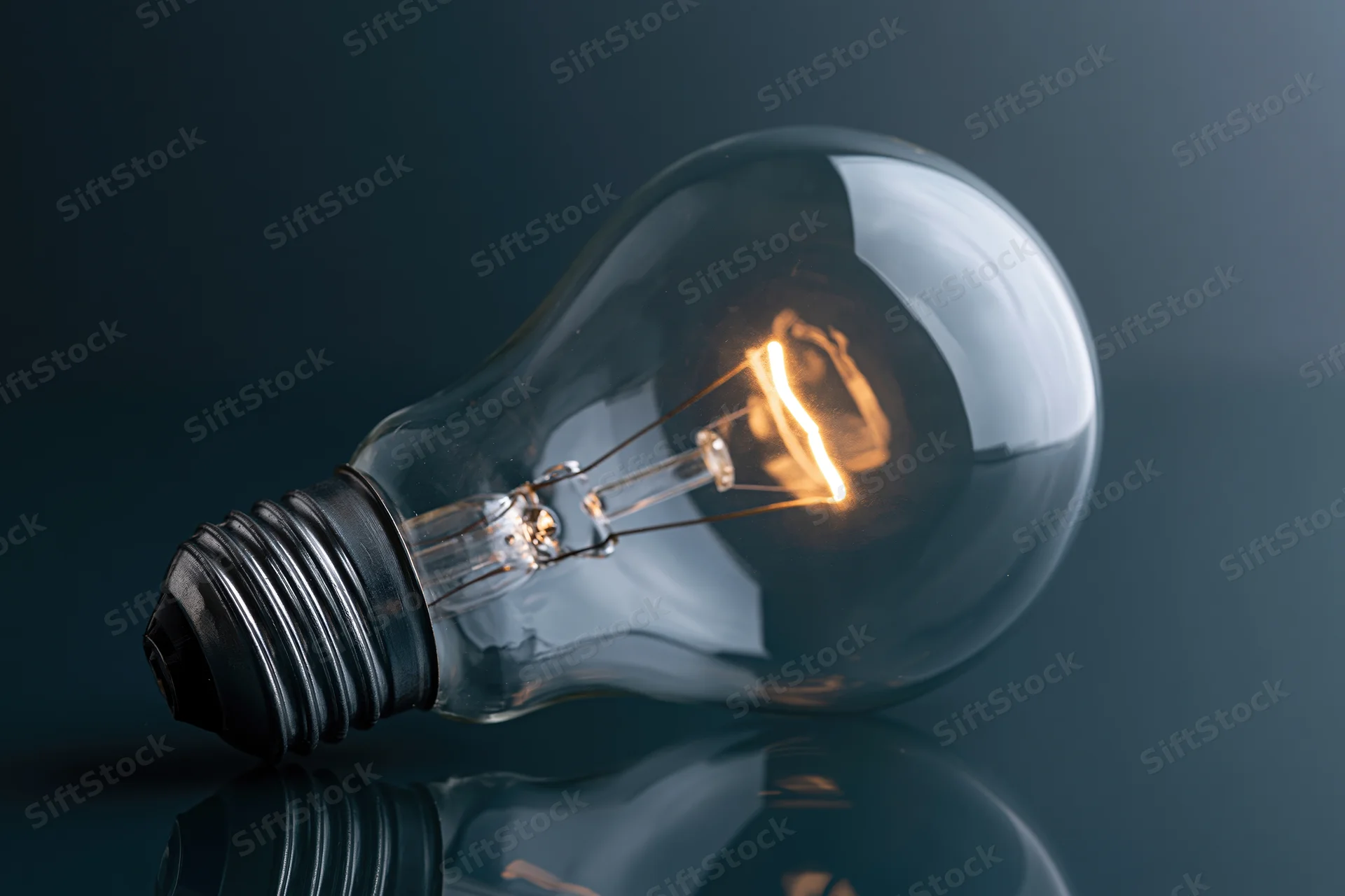 Illuminated incandescent light bulb lying on its side on a reflective dark surface