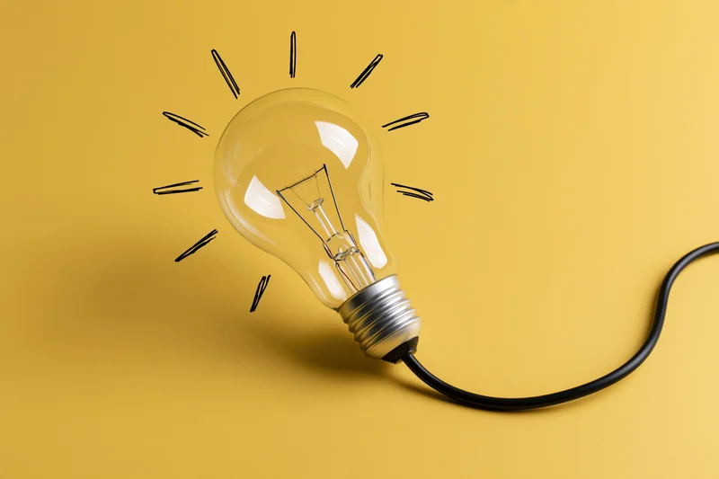 Clear glass incandescent light bulb with hand-drawn rays on yellow background, connected by black cord