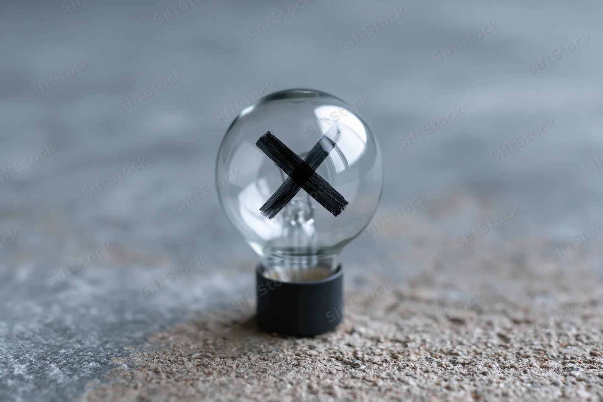 Clear glass lightbulb with black x painted inside resting on textured gray surface