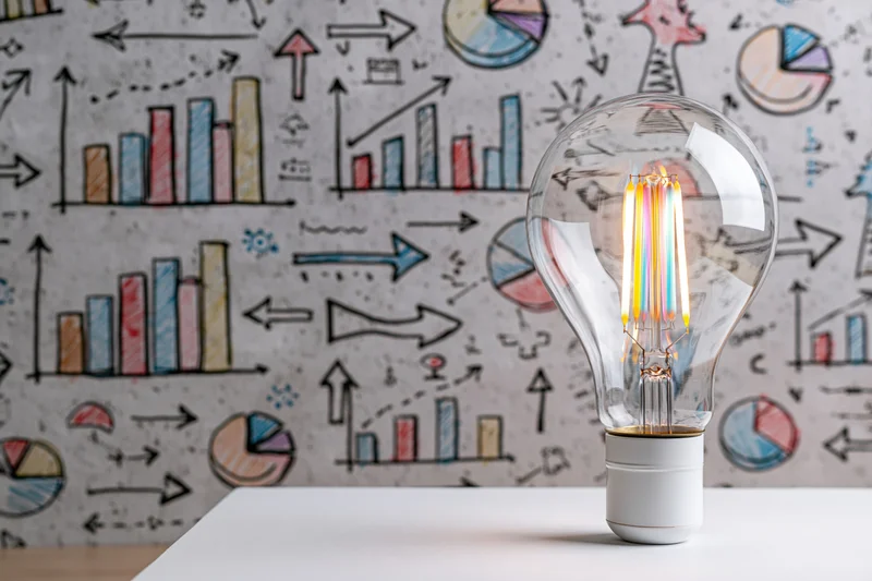 Glowing multicolored filament lightbulb on white surface with hand-drawn business charts and graphs in background