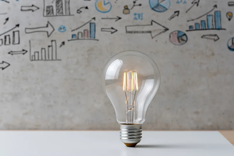 Glowing lightbulb on a desk with hand-drawn business diagrams on a concrete wall behind it