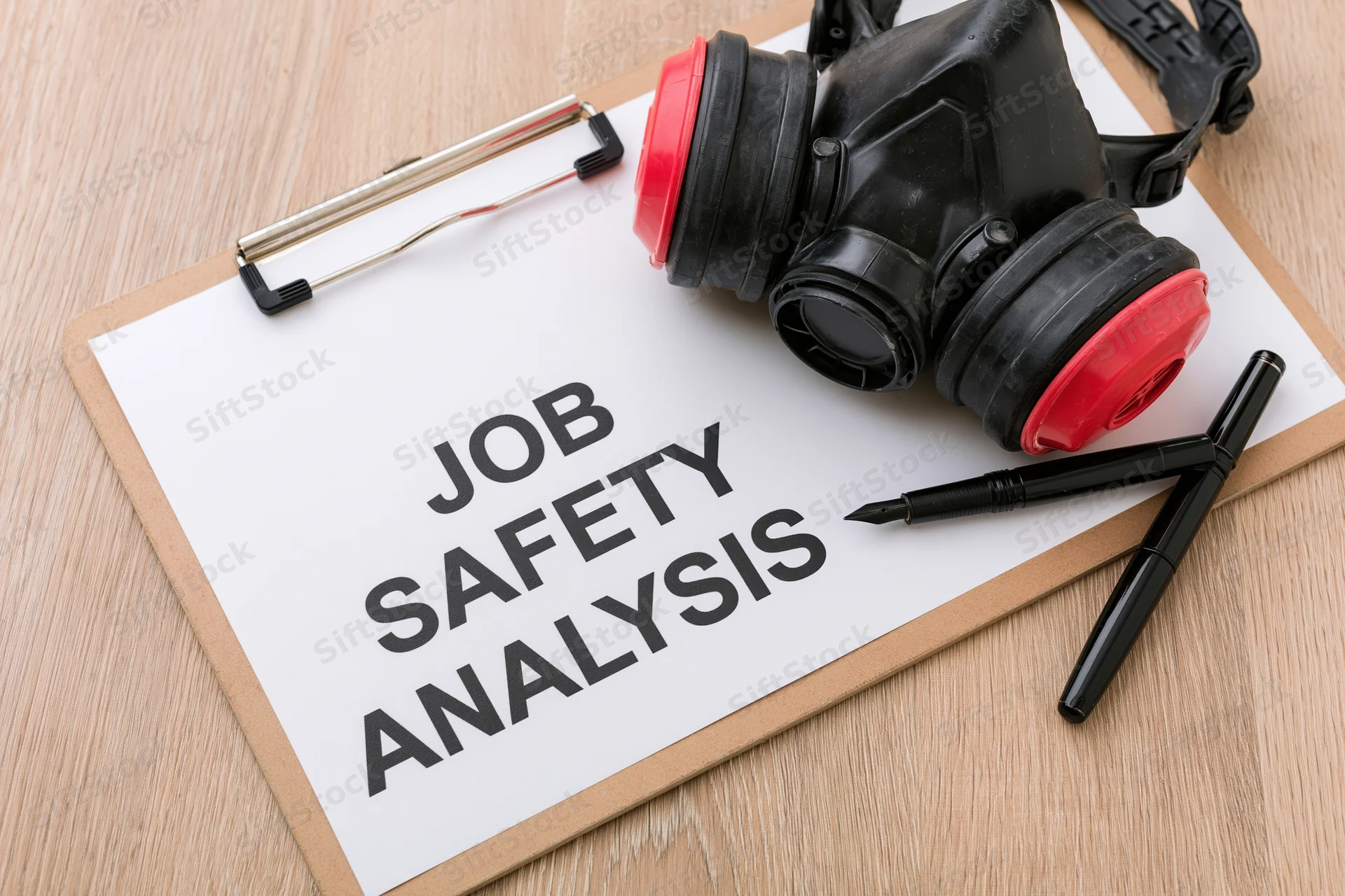 Clipboard with paper labeled job safety analysis and respirator mask on wooden surface