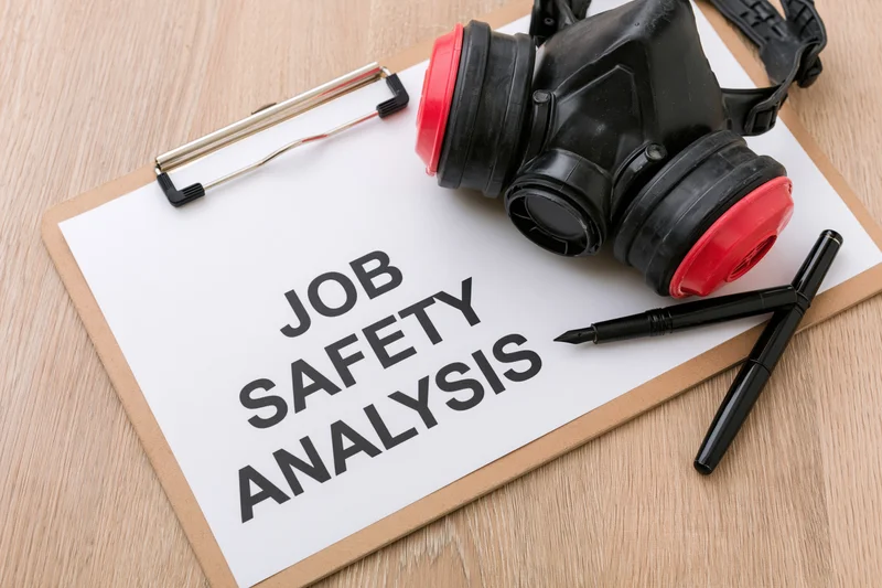 Clipboard with paper labeled job safety analysis and respirator mask on wooden surface