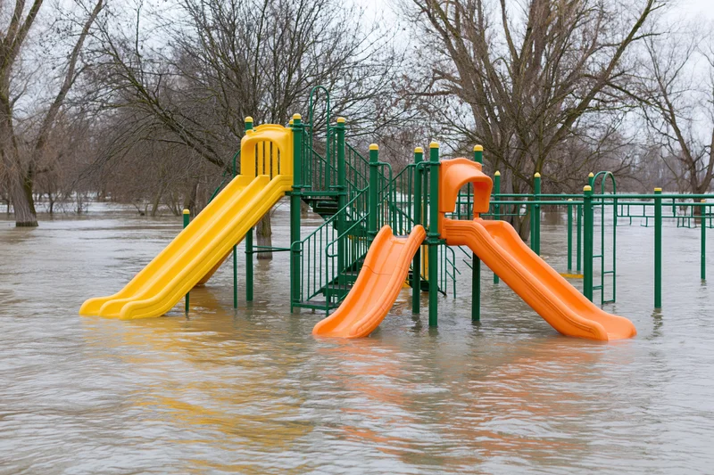 Colorful childrens playground structure partially submerged in murky floodwater with bare trees in background