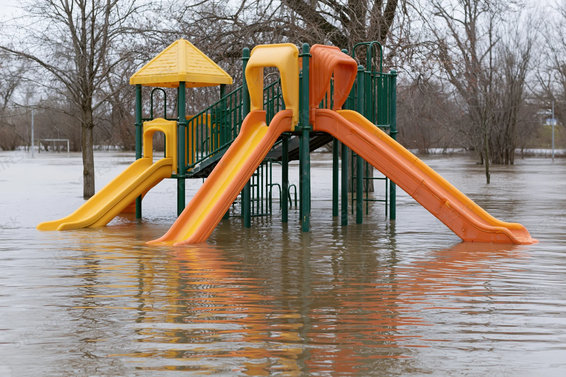 Colorful childrens playground structure partially submerged in murky floodwater with bare trees in background