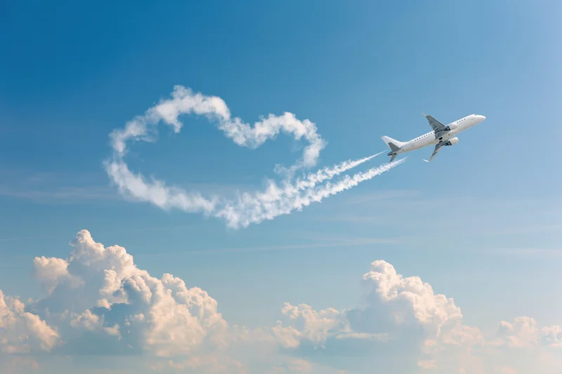 Commercial airplane climbing through open sky leaving heart-shaped contrail in blue sky with cumulus clouds below