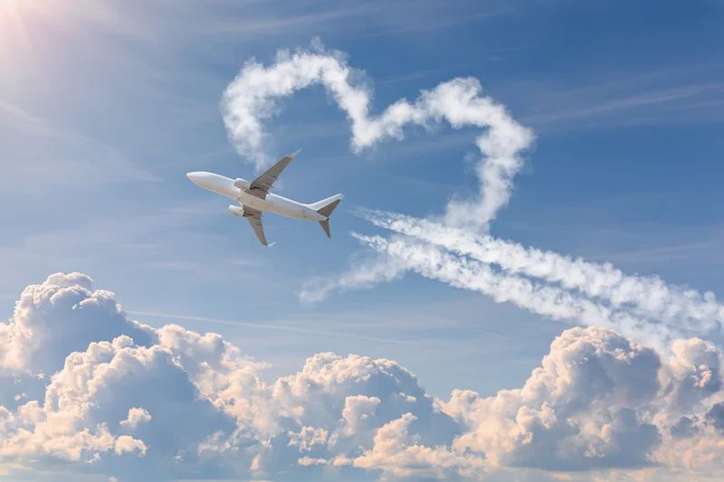 Commercial airplane climbing through sky with heart-shaped cloud formation above fluffy cumulus clouds