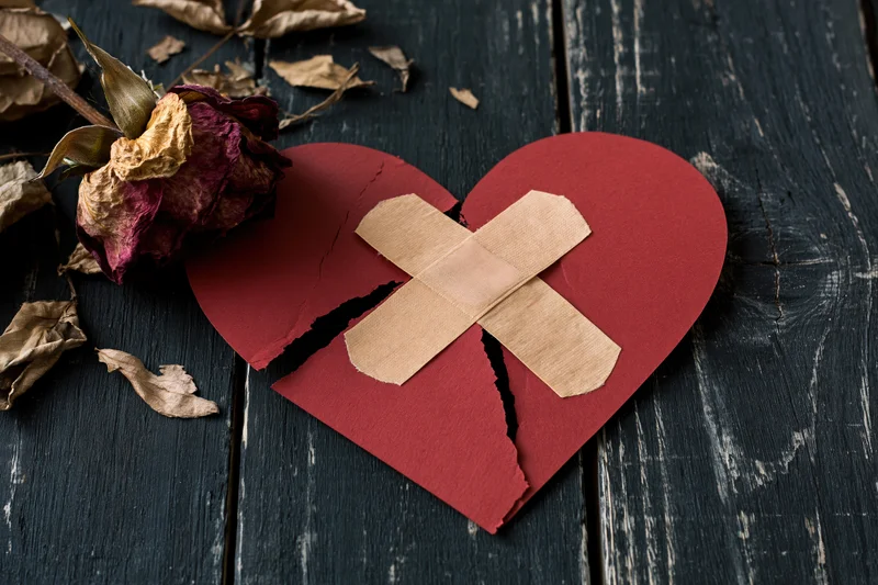 Broken red paper heart with crossed tape bandage on dark wooden surface beside dried rose petals