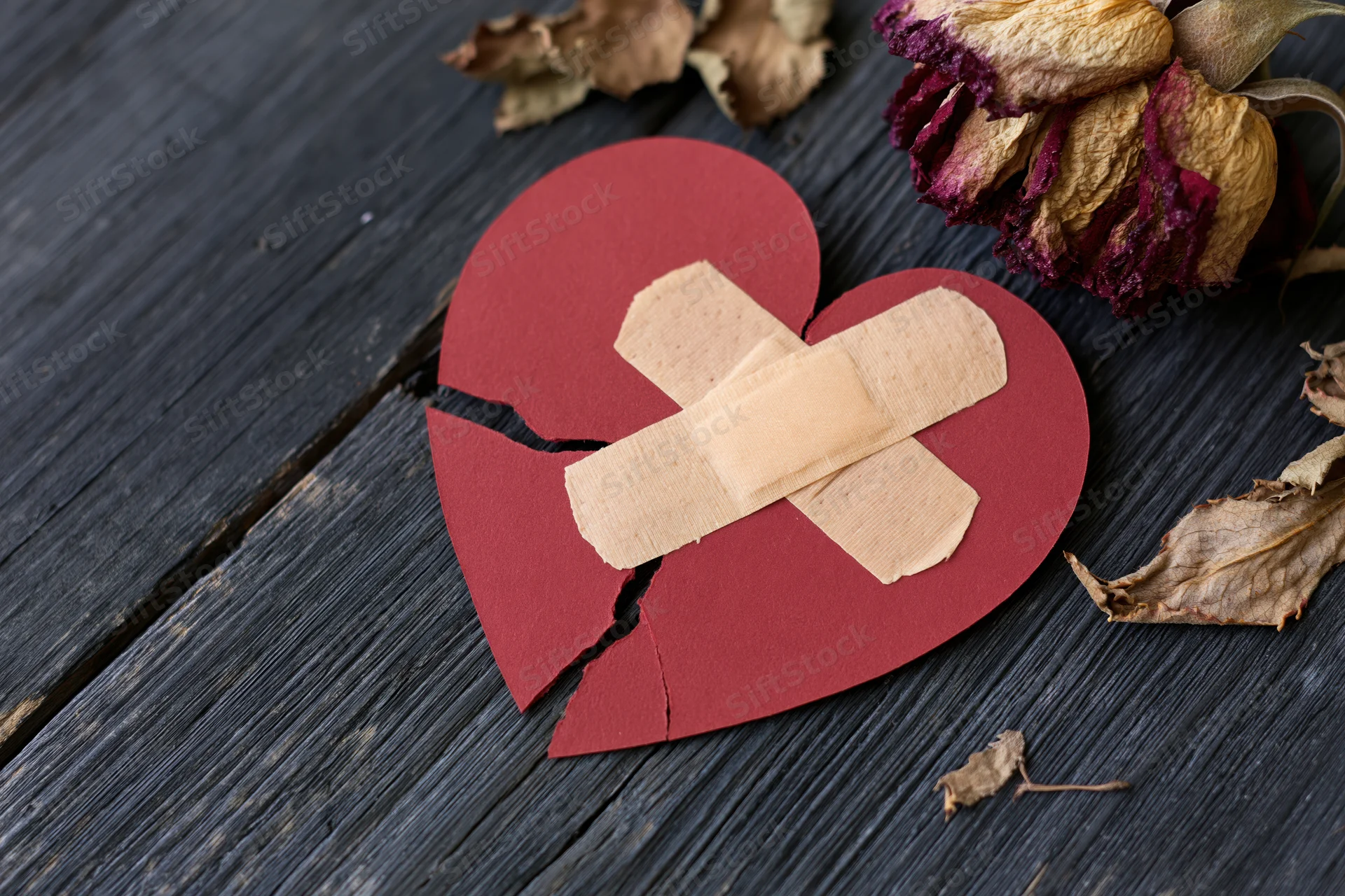 Broken red paper heart with crossed bandages on dark wooden surface beside dried roses