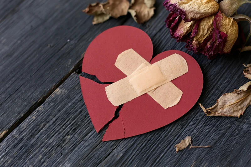 Broken red paper heart with crossed bandages on dark wooden surface beside dried roses