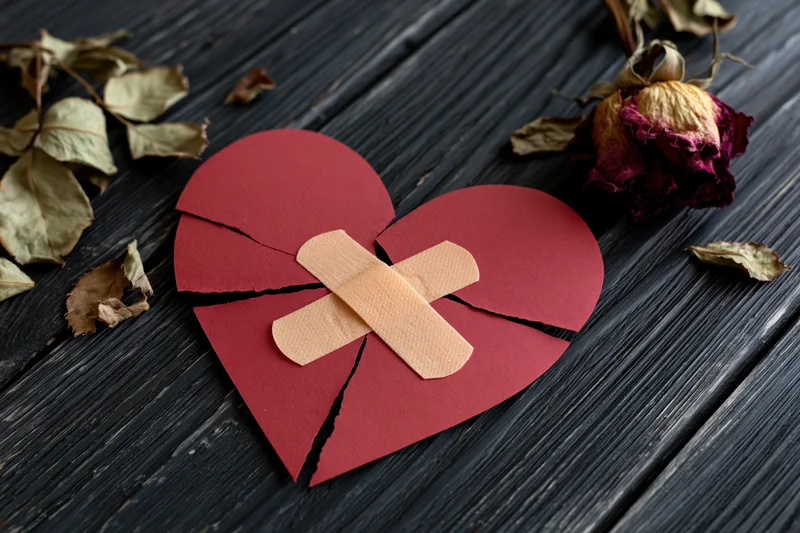 Broken red paper heart held together with crossed bandages on dark wooden surface surrounded by dried rose petals