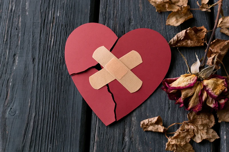 Cracked red paper heart with crossed bandage on dark wood beside dried rose petals