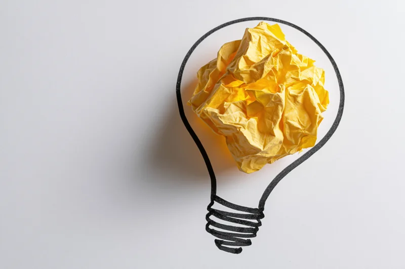 Crumpled yellow paper ball inside hand-drawn lightbulb outline on white surface