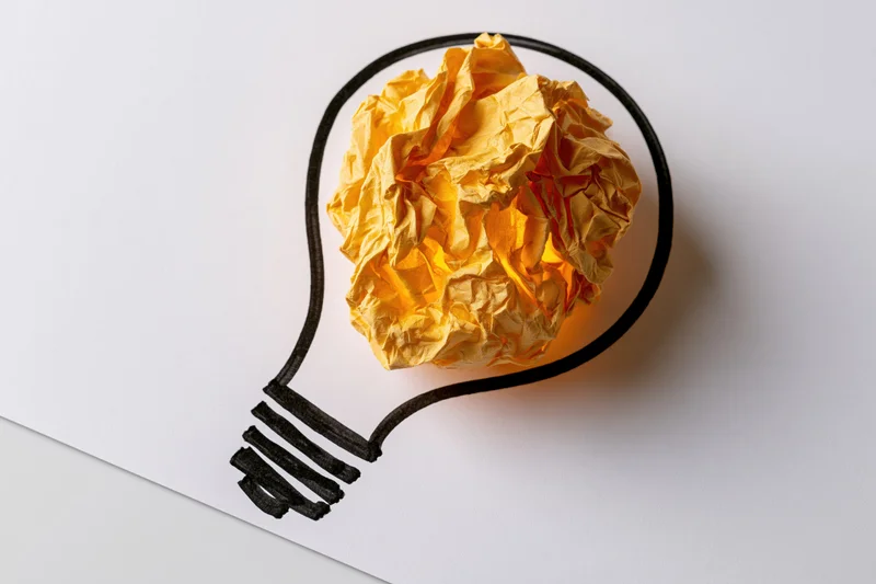 Crumpled yellow paper ball inside hand-drawn lightbulb outline on white paper