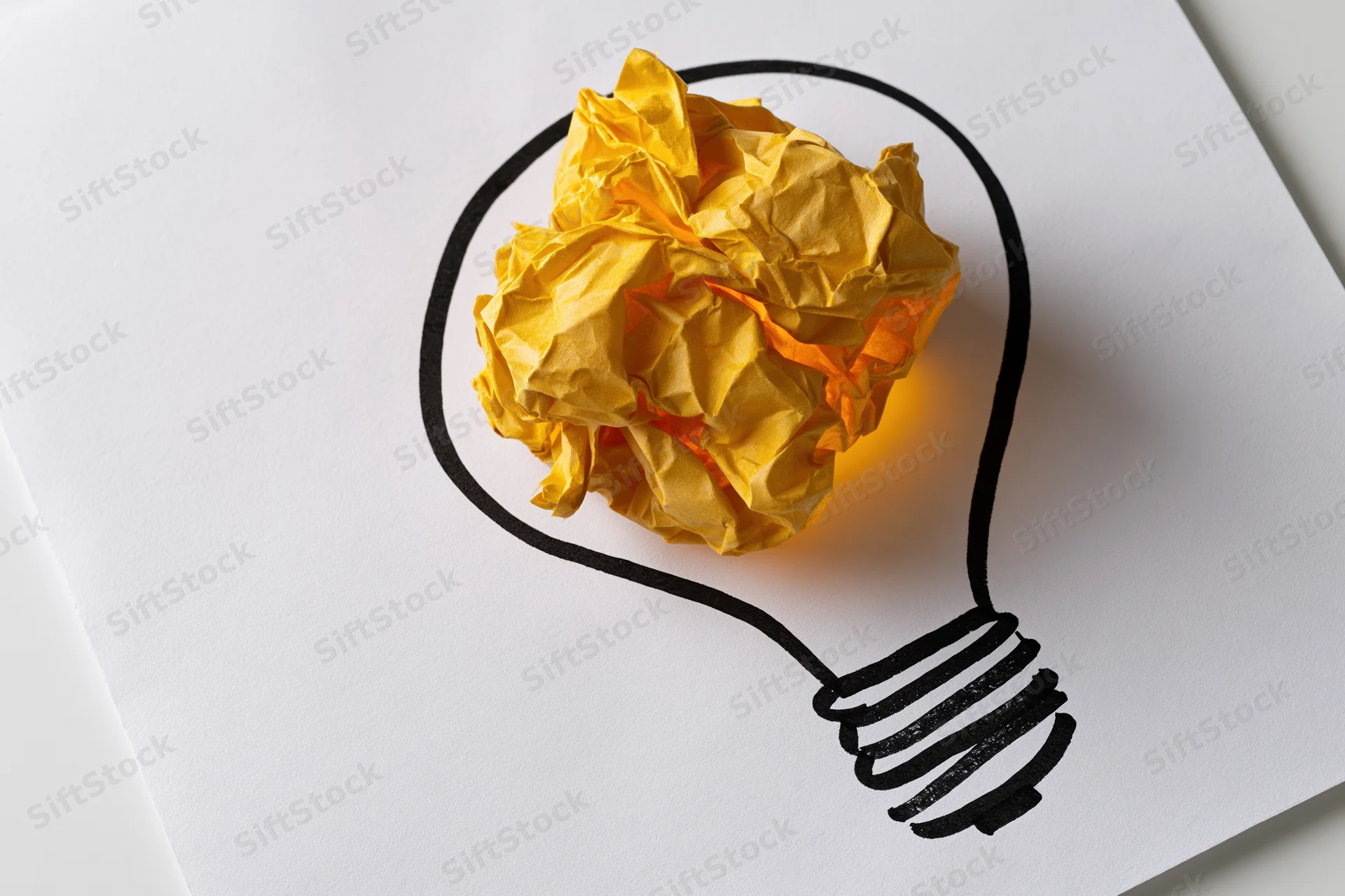 Crumpled yellow paper ball glowing inside hand-drawn lightbulb outline on white sheet