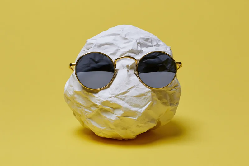 Crumpled white paper ball wearing round gold-frame sunglasses on yellow background