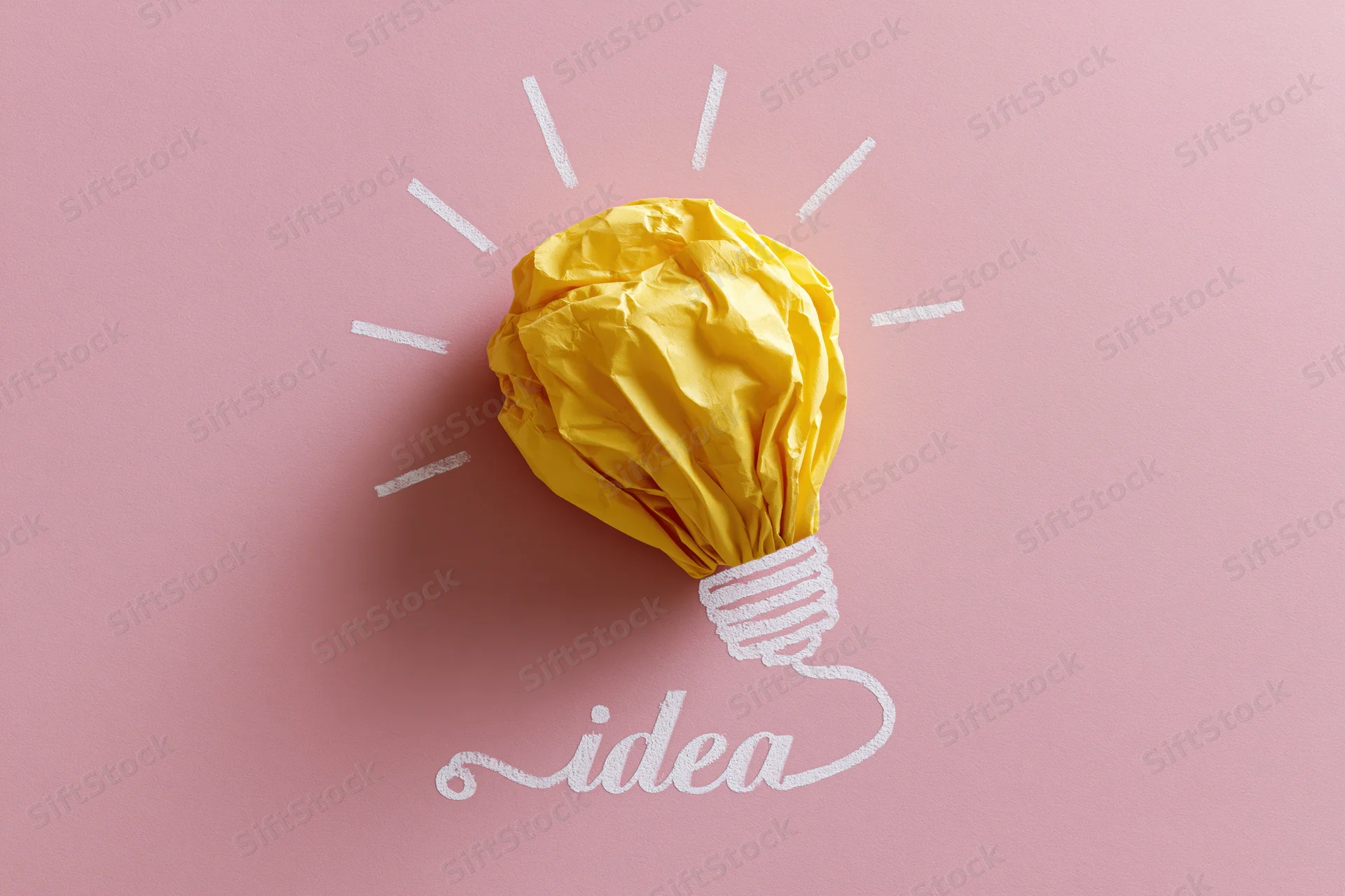 Crumpled yellow paper ball shaped as a light bulb on pink background with white rays and idea text
