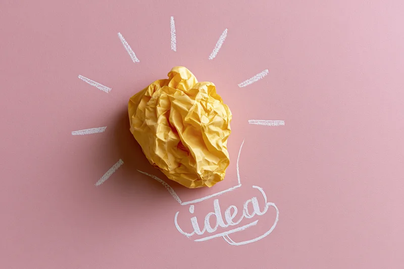 Crumpled yellow paper ball arranged as a light bulb on pink background with white sketch lines and word idea
