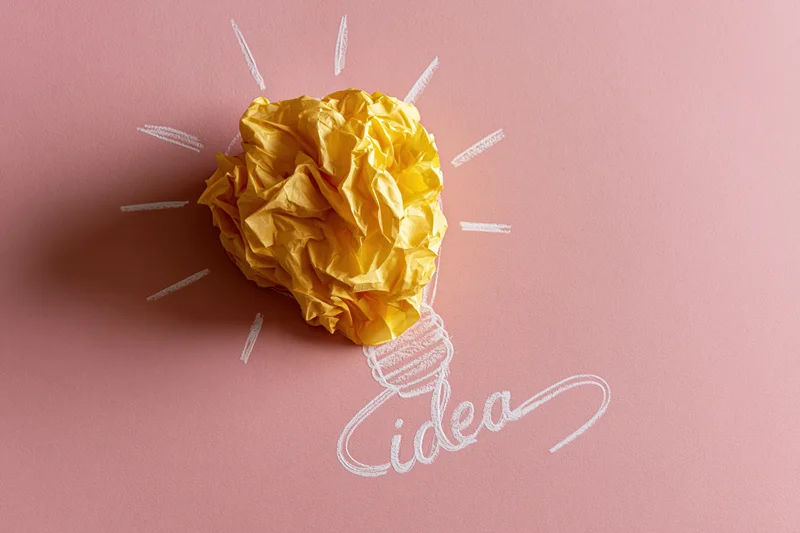 Crumpled yellow paper ball shaped as a light bulb with chalk-drawn rays and the word idea on a pink surface
