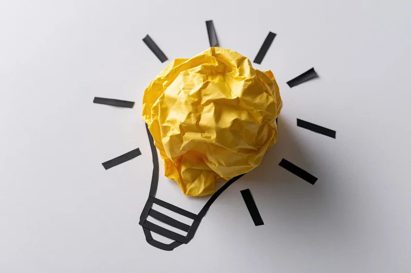 Crumpled yellow paper ball forming a lightbulb shape on white surface with black lines radiating from it
