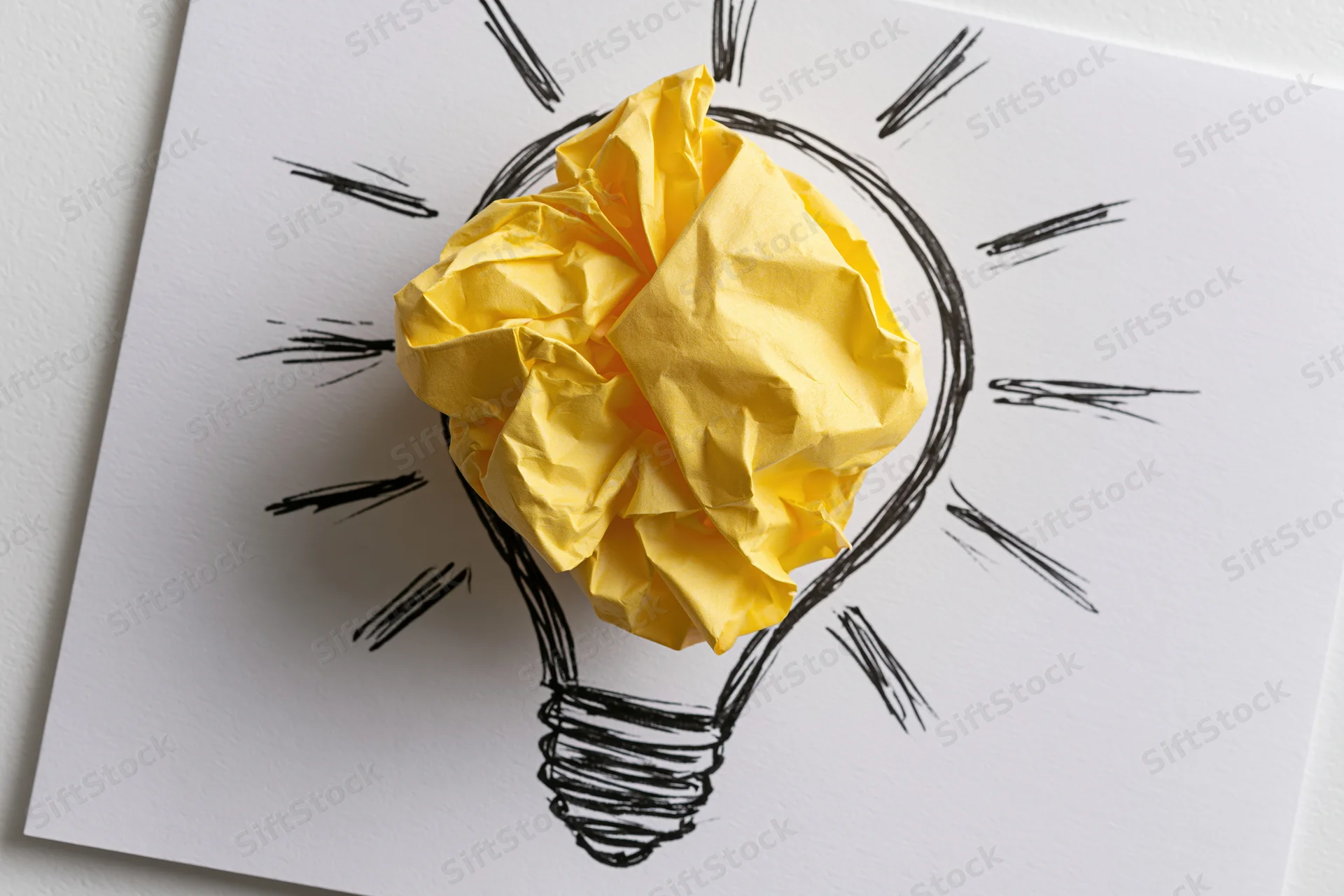 Crumpled yellow paper ball placed inside hand-drawn lightbulb sketch on white paper
