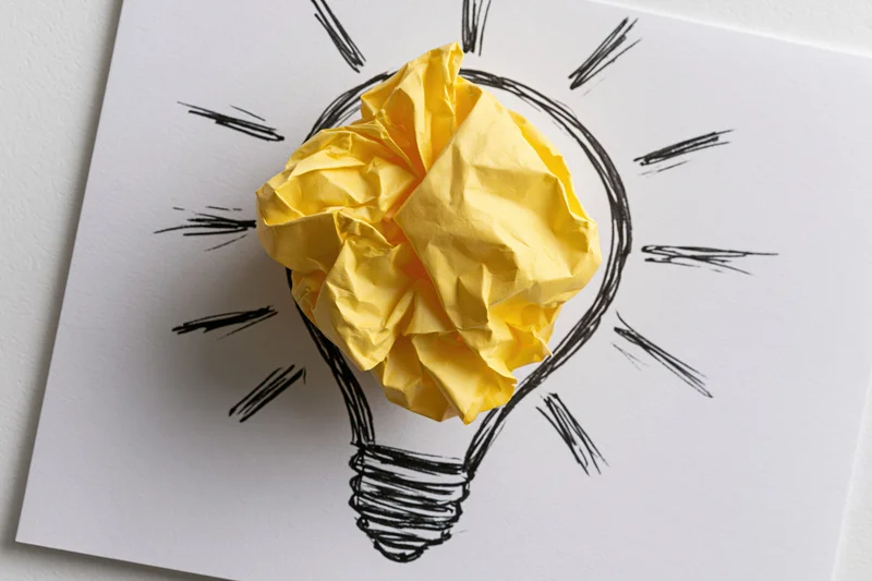 Crumpled yellow paper ball placed inside hand-drawn lightbulb sketch on white paper