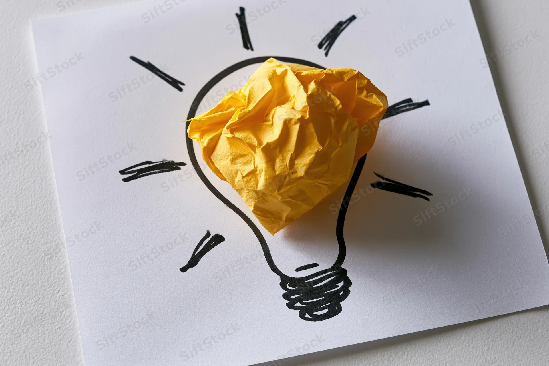Crumpled yellow paper ball placed inside hand-drawn lightbulb on white paper sheet