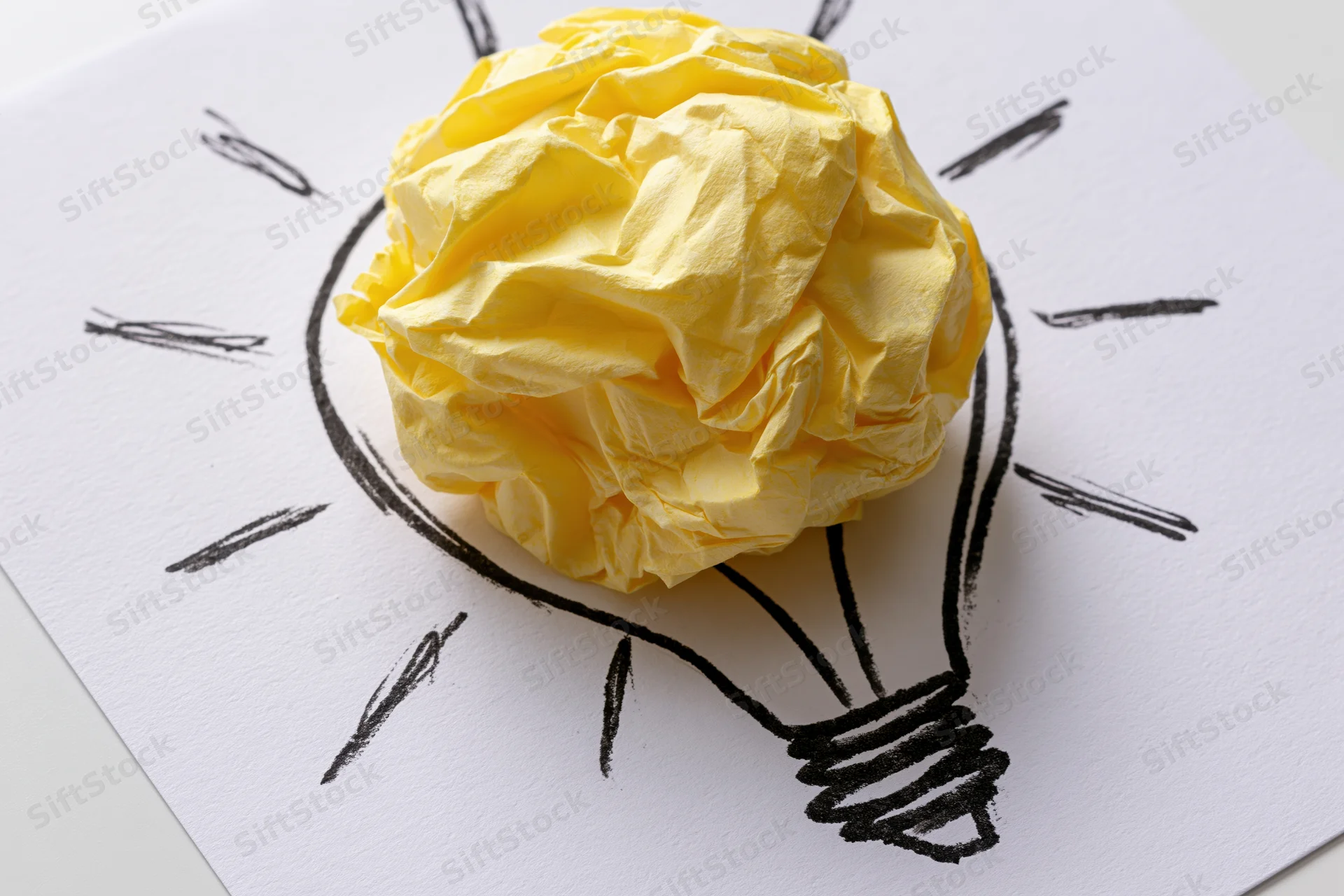 Crumpled yellow paper ball placed inside hand-drawn lightbulb outline on white paper