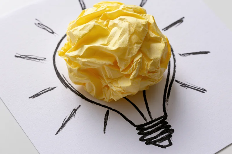 Crumpled yellow paper ball placed inside hand-drawn lightbulb outline on white paper