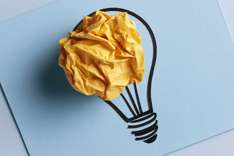 Crumpled yellow paper ball placed inside hand-drawn lightbulb on blue paper