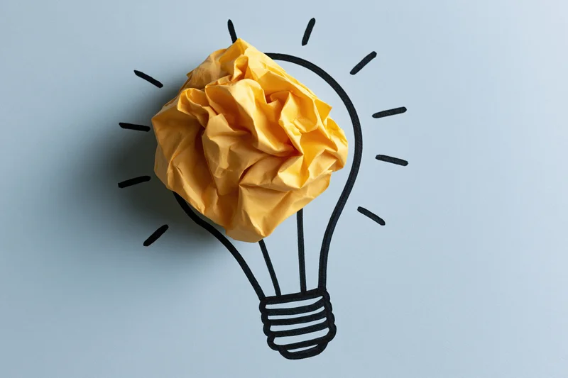 Crumpled yellow paper ball placed inside hand-drawn lightbulb outline on light blue background