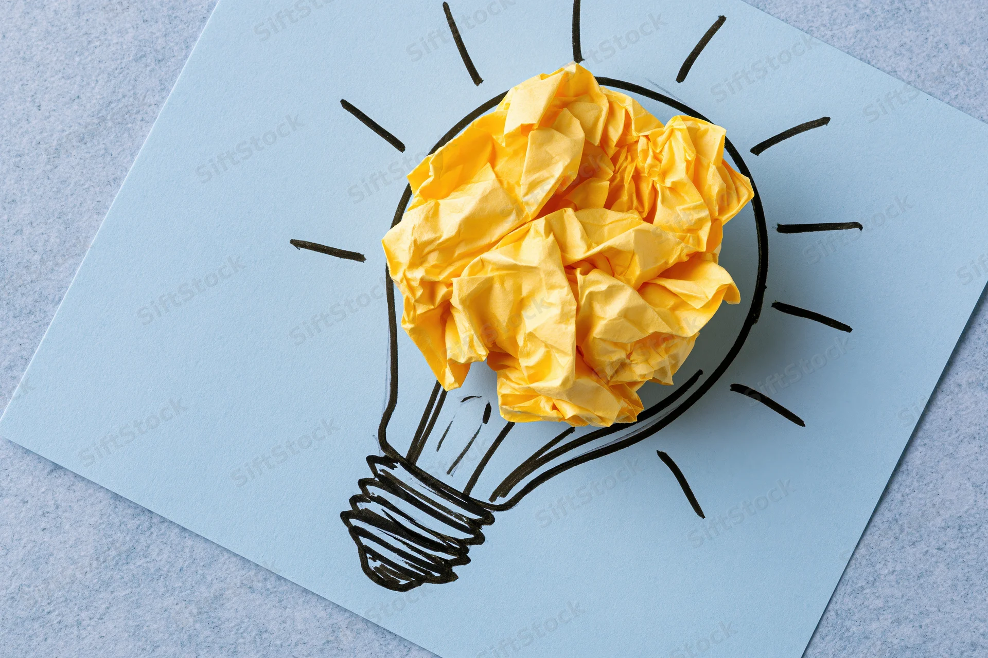 Crumpled yellow paper ball placed inside hand-drawn lightbulb on blue paper