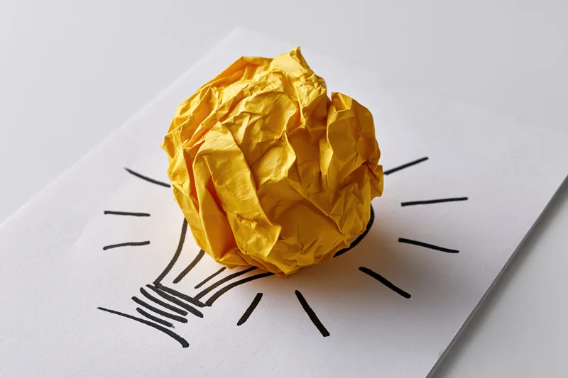 Crumpled yellow paper ball placed on a hand-drawn lightbulb sketch on white paper