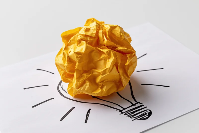 Crumpled yellow paper ball placed on a hand-drawn lightbulb sketch on white paper