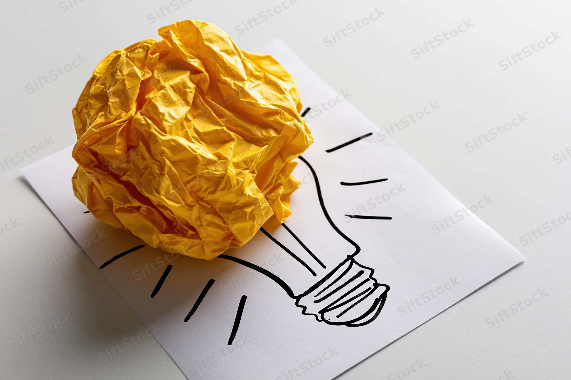 Crumpled yellow paper ball placed on a hand-drawn lightbulb sketch on white paper