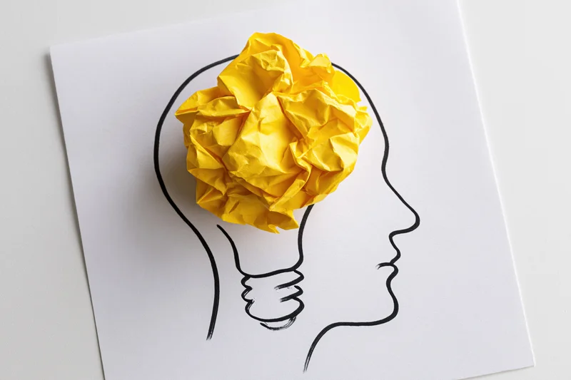 Crumpled yellow paper ball inside a hand-drawn lightbulb head outline on white paper