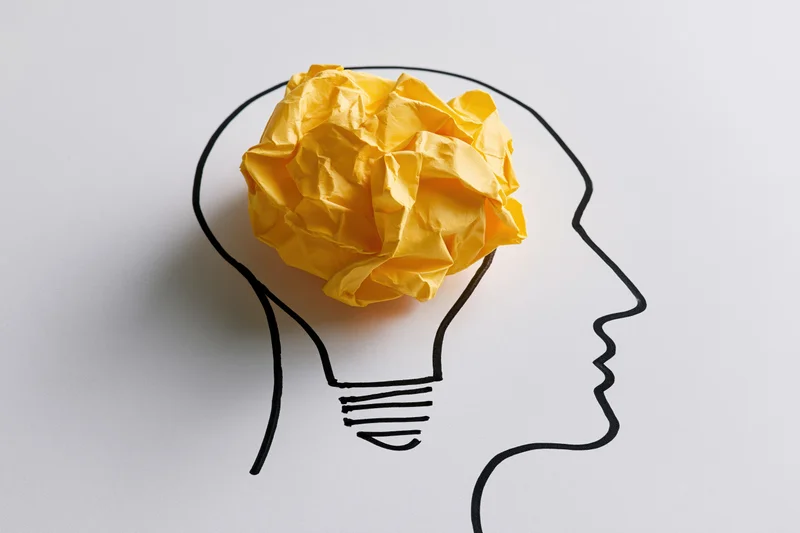 Crumpled yellow paper ball inside a drawn lightbulb-shaped head outline on white background