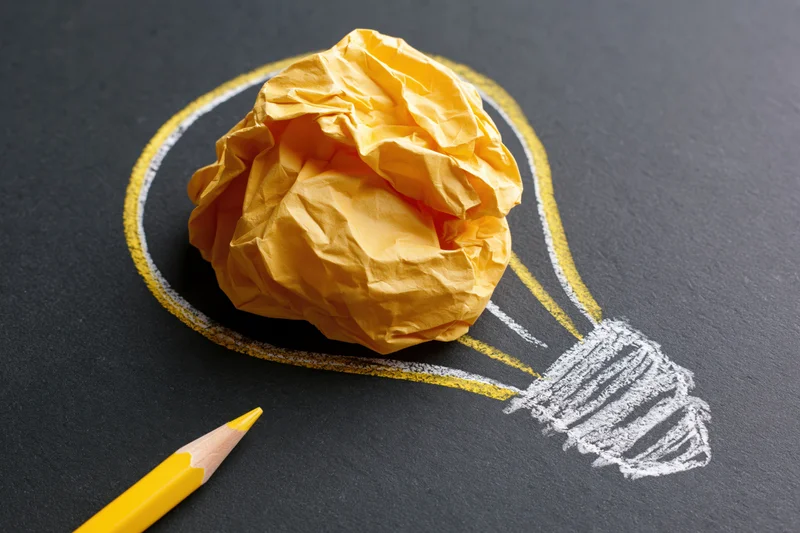 Crumpled yellow paper ball inside a chalk-drawn lightbulb on dark surface with yellow pencil nearby