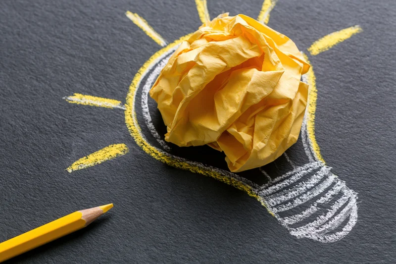 Crumpled yellow paper ball placed inside a chalk-drawn lightbulb on dark surface with yellow pencil nearby