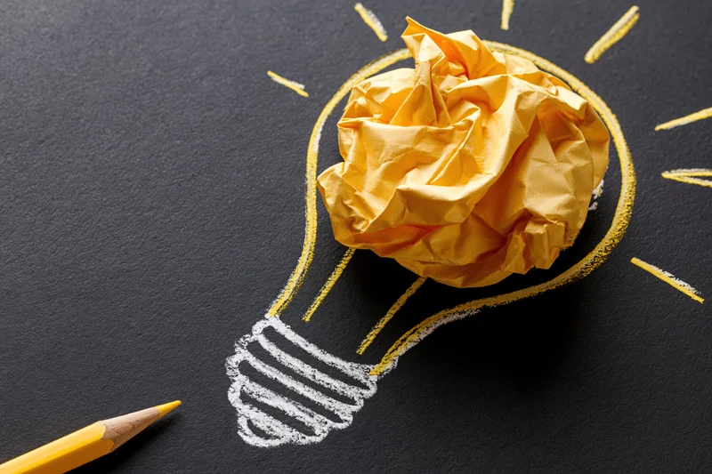 Crumpled yellow paper ball placed inside a hand-drawn lightbulb outline on black surface with yellow pencil nearby