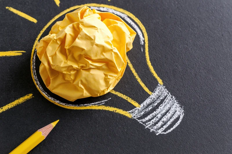 Crumpled yellow paper ball inside hand-drawn lightbulb on blackboard with yellow pencil nearby