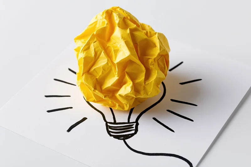 Crumpled yellow paper ball placed on a hand-drawn lightbulb sketch on white paper